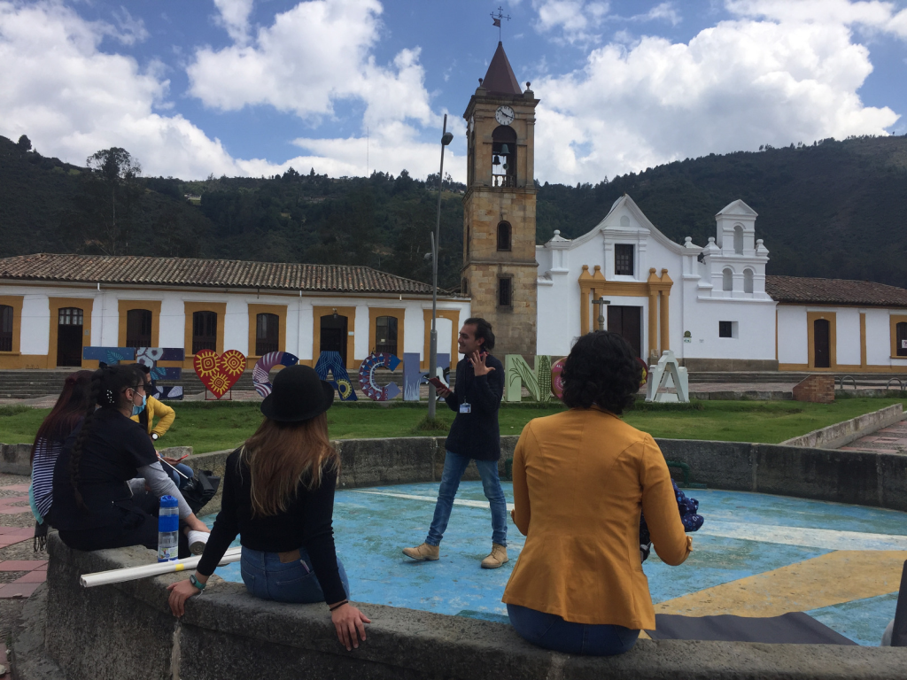 Plaza del municipio de Gachancipá, al fondo iglesia y edificación colonial. Una joven de pie habla a otras licenciadas en formación sentadas alrededor. 