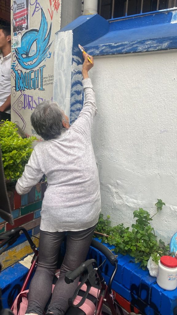 Señora adulta mayor con brocha en la mano, pinta una especie de nubes blancas sobre fondo azul que enmarca mural de Aula Húmeda. 