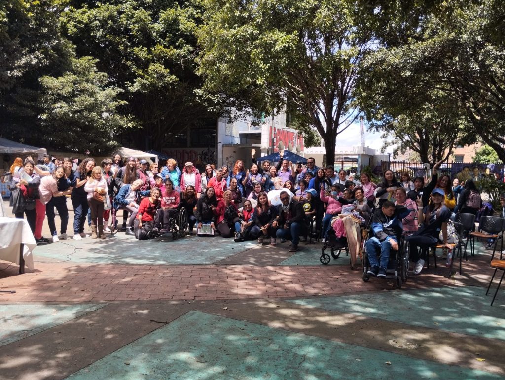 Grupo de adultos y jóvenes participantes de Aula Húmeda, en plaza frente al edificio C. En el grupo personas ciegas y usuarias de silla de ruedas entre otras condiciones diversas. 